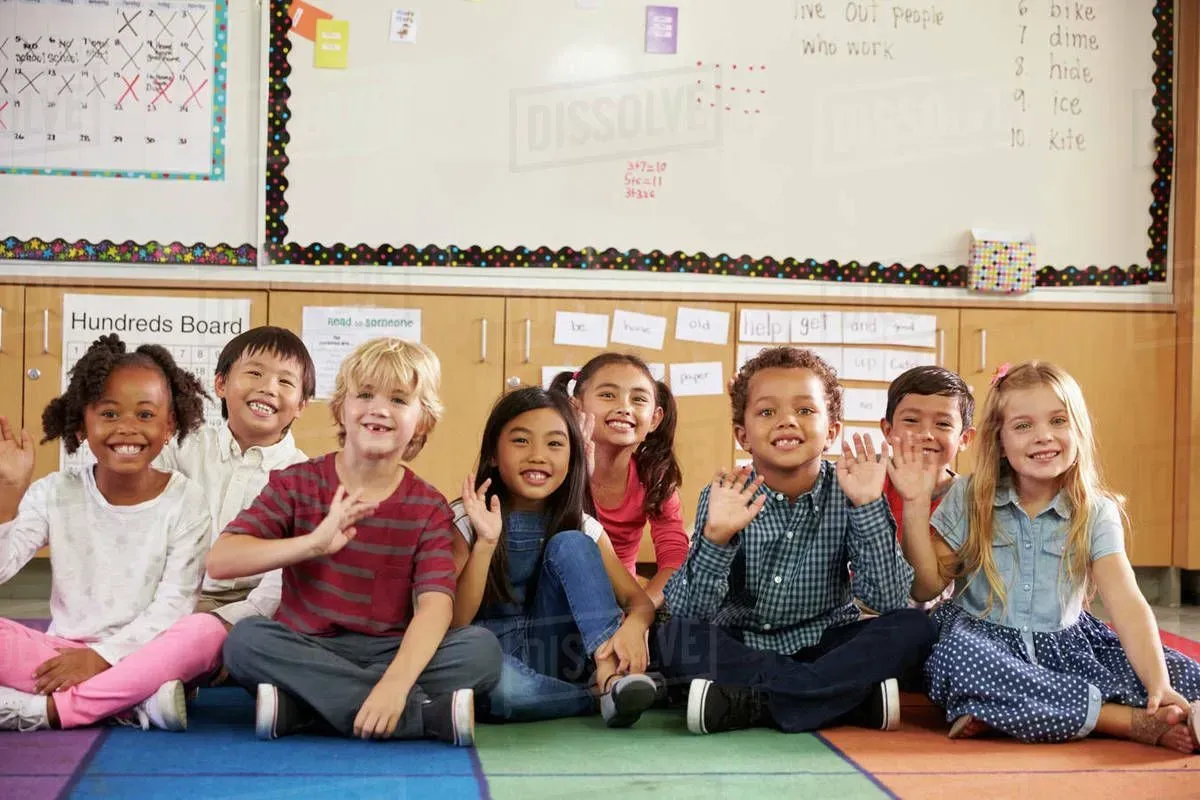 Group of children sitting on a colorful floor in a classroom, waving.