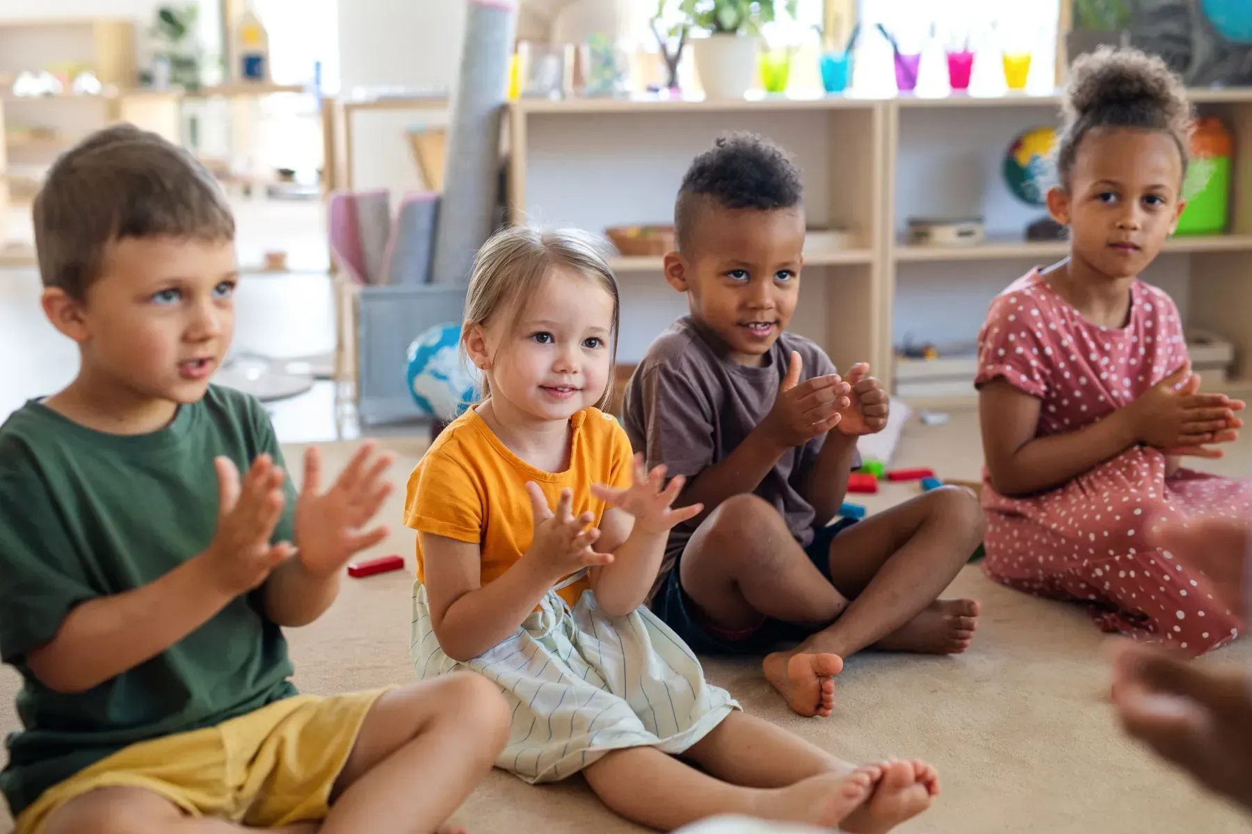 Four children sitting, clapping. Brightly lit room, shelves in background.