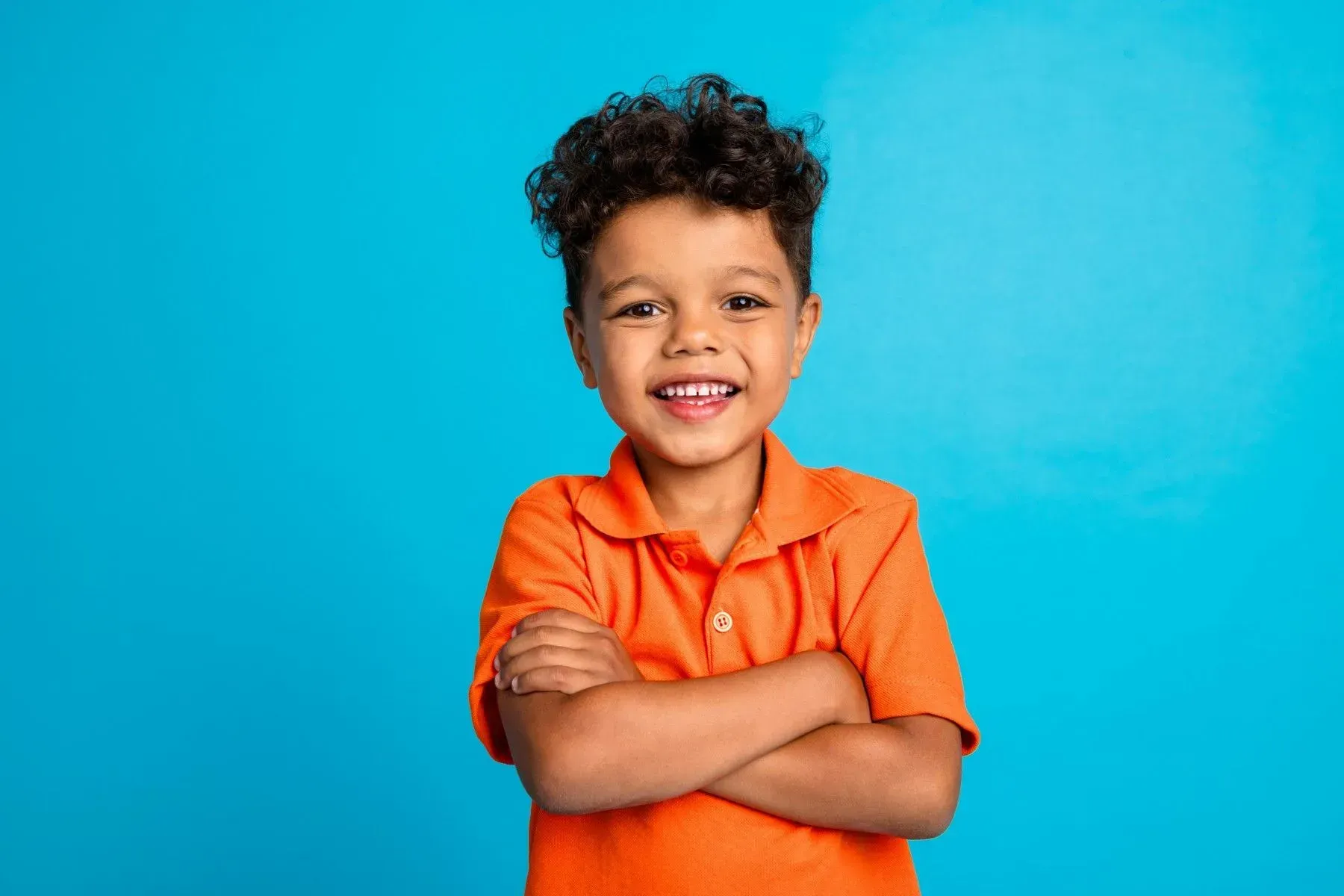 Boy with arms crossed, smiling, wearing an orange shirt against a blue background.