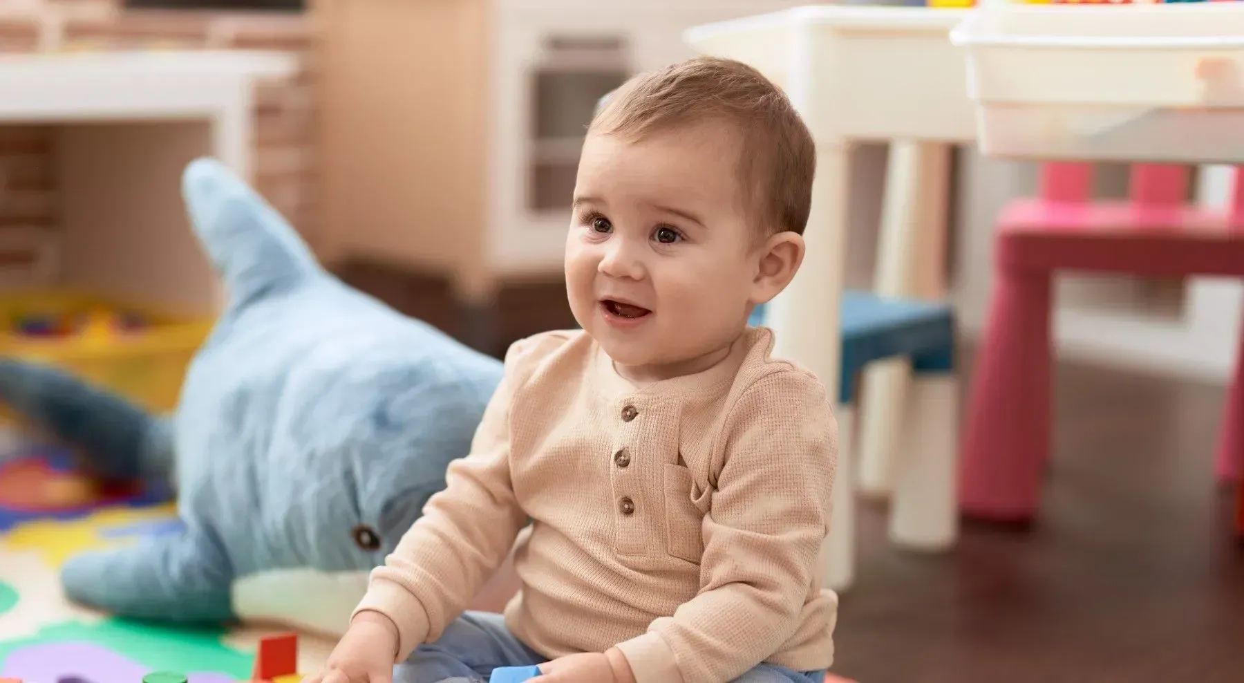 Smiling baby sitting in a playroom, near a plush shark and small table.