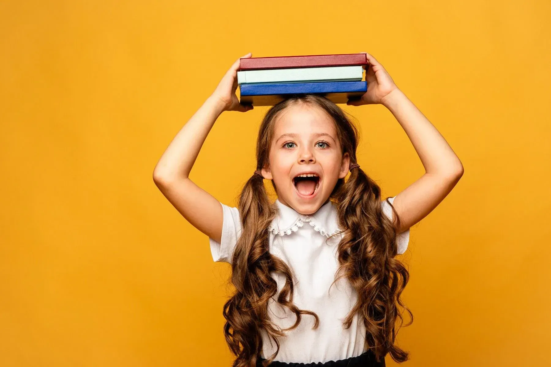 Girl with long brown hair, holding a stack of books on her head, excited expression.