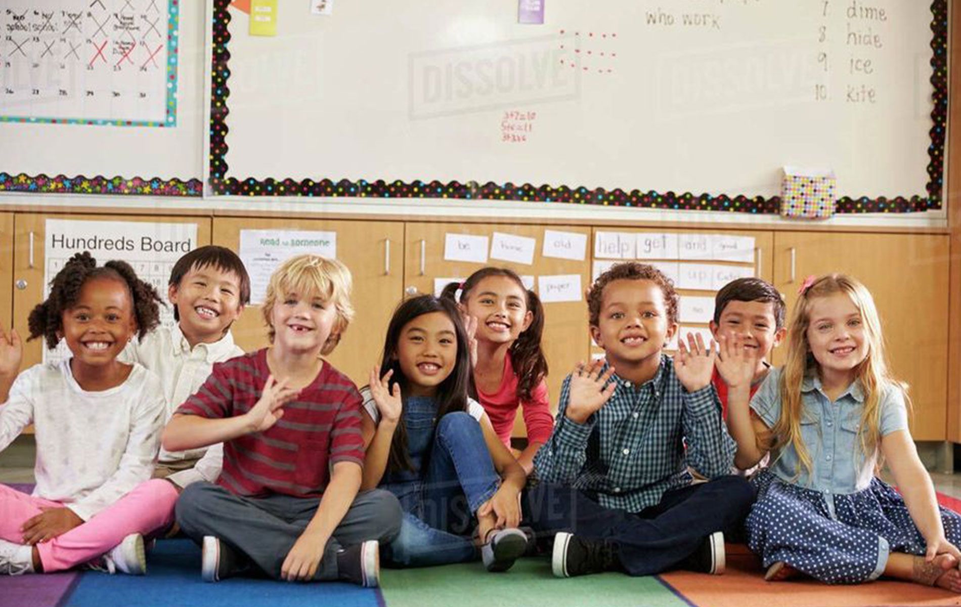 Group of children sitting and waving in a classroom.
