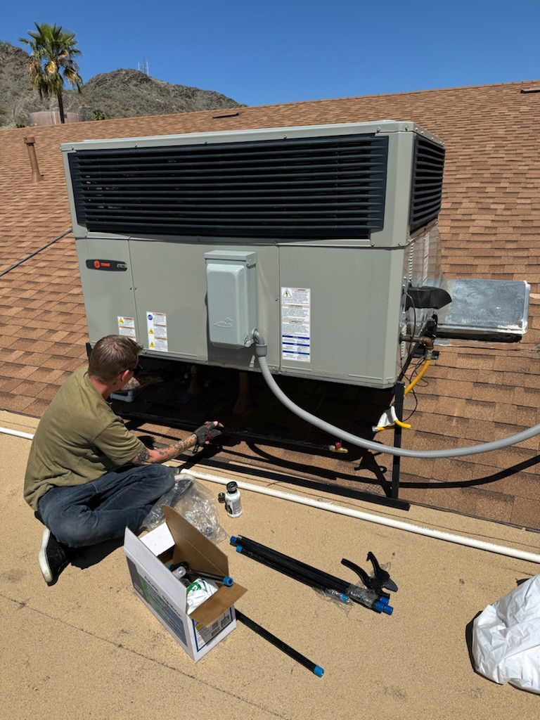 A man is kneeling on the ground working on an air conditioner on top of a roof.