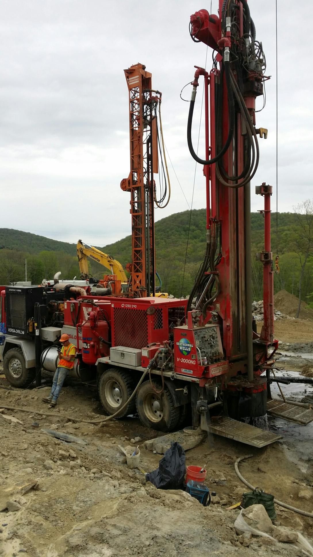 Two workers operating a drilling rig, one looking up, water spraying, grassy field.