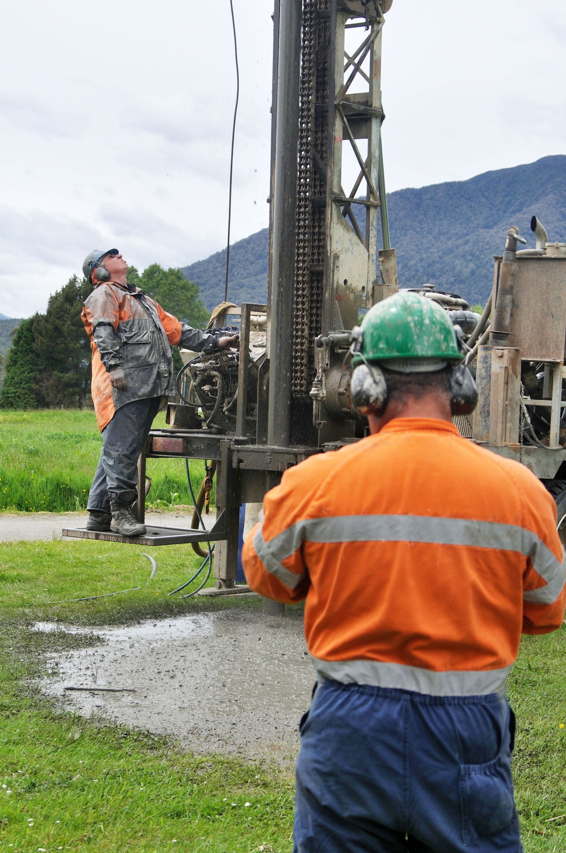 Two workers operating a drilling rig, one looking up, water spraying, grassy field.