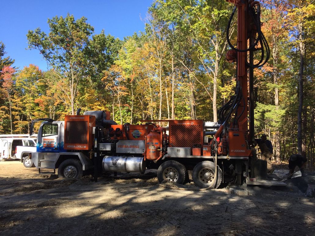 Blue water pump with large pulley, connected to blue pipes, set outdoors among trees.