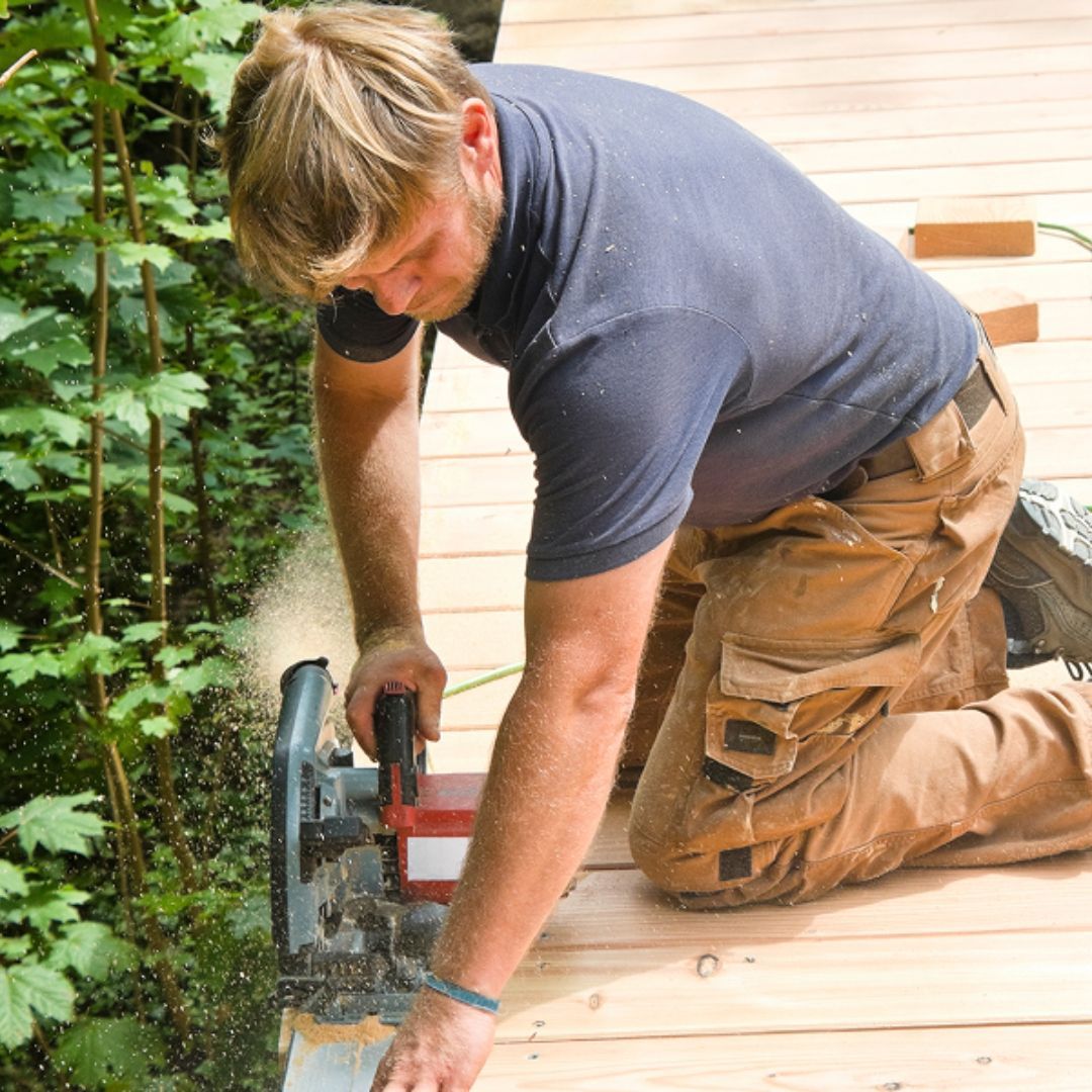 A person in a blue polo and tan work pants kneels on a wooden deck, using a circular saw to cut a board.