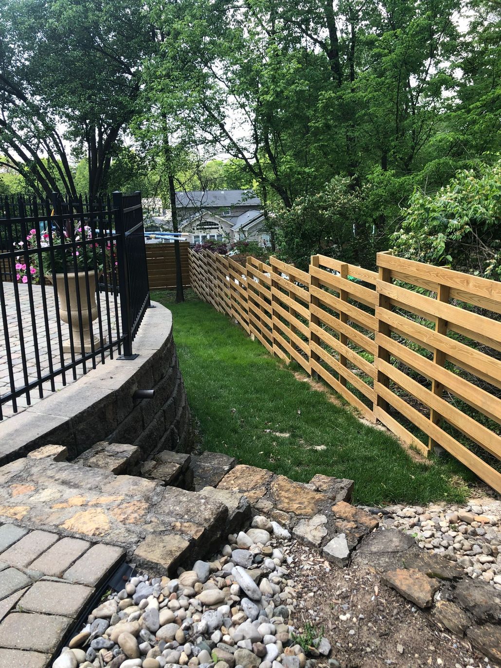Walkway beside a black fence and wooden fence, with rocks, grass, and trees in a yard