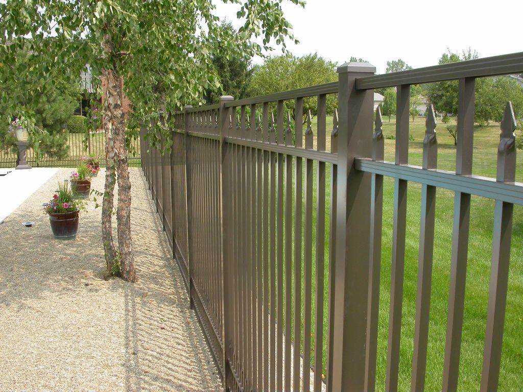 Brown metal fence beside a gravel path with potted flowers and trees in a sunny yard