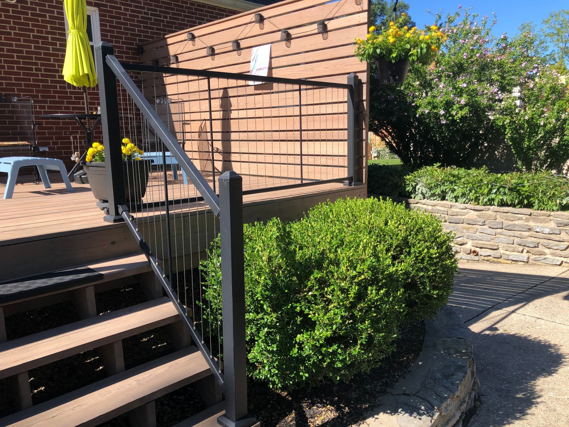 Outdoor stairs beside a brick house, with a black railing and neatly trimmed bushes in a sunny yard.