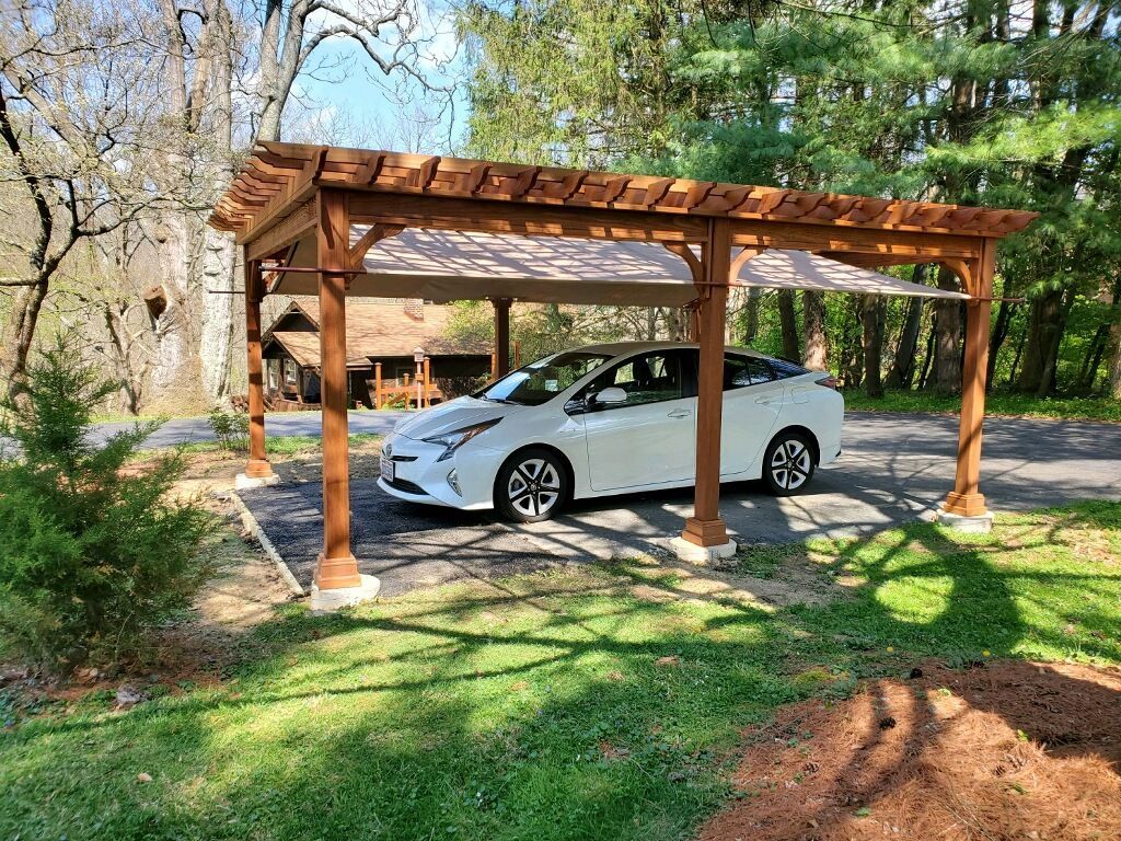 White sedan parked under a wooden carport in a wooded yard with green grass and trees