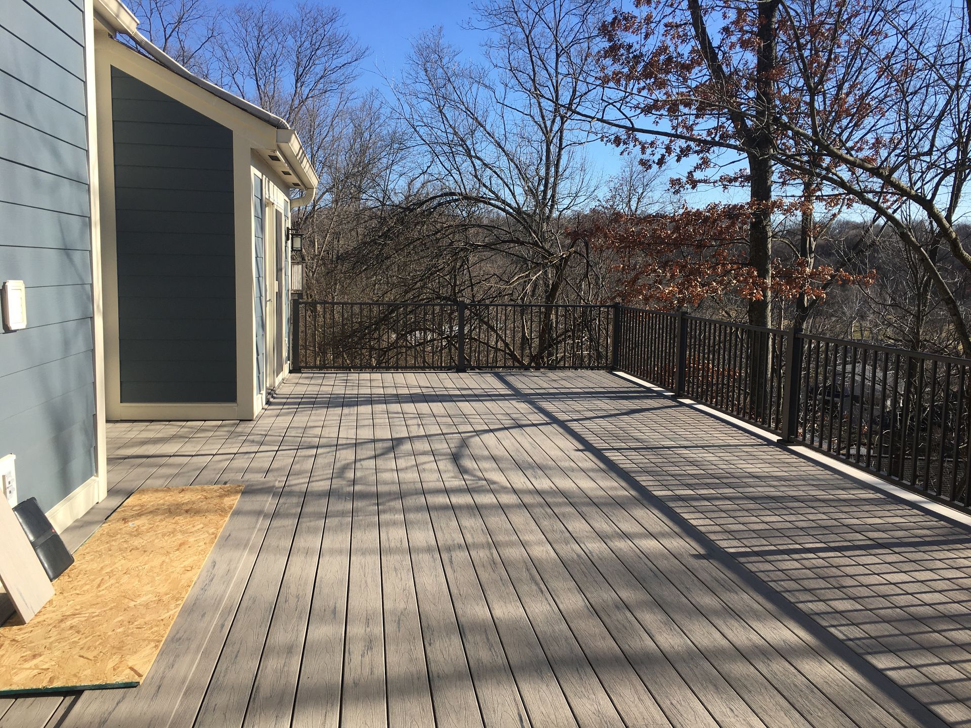 Wide wooden deck beside a house, bordered by black railings and leafless trees in bright sunlight