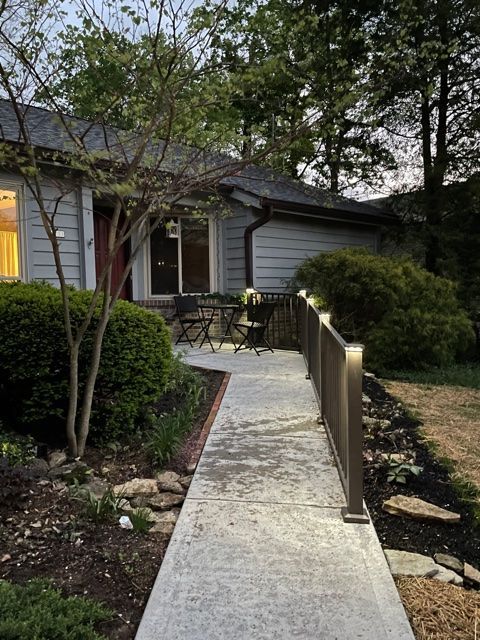 Concrete walkway with handrails leading to a house porch at dusk, surrounded by shrubs and trees