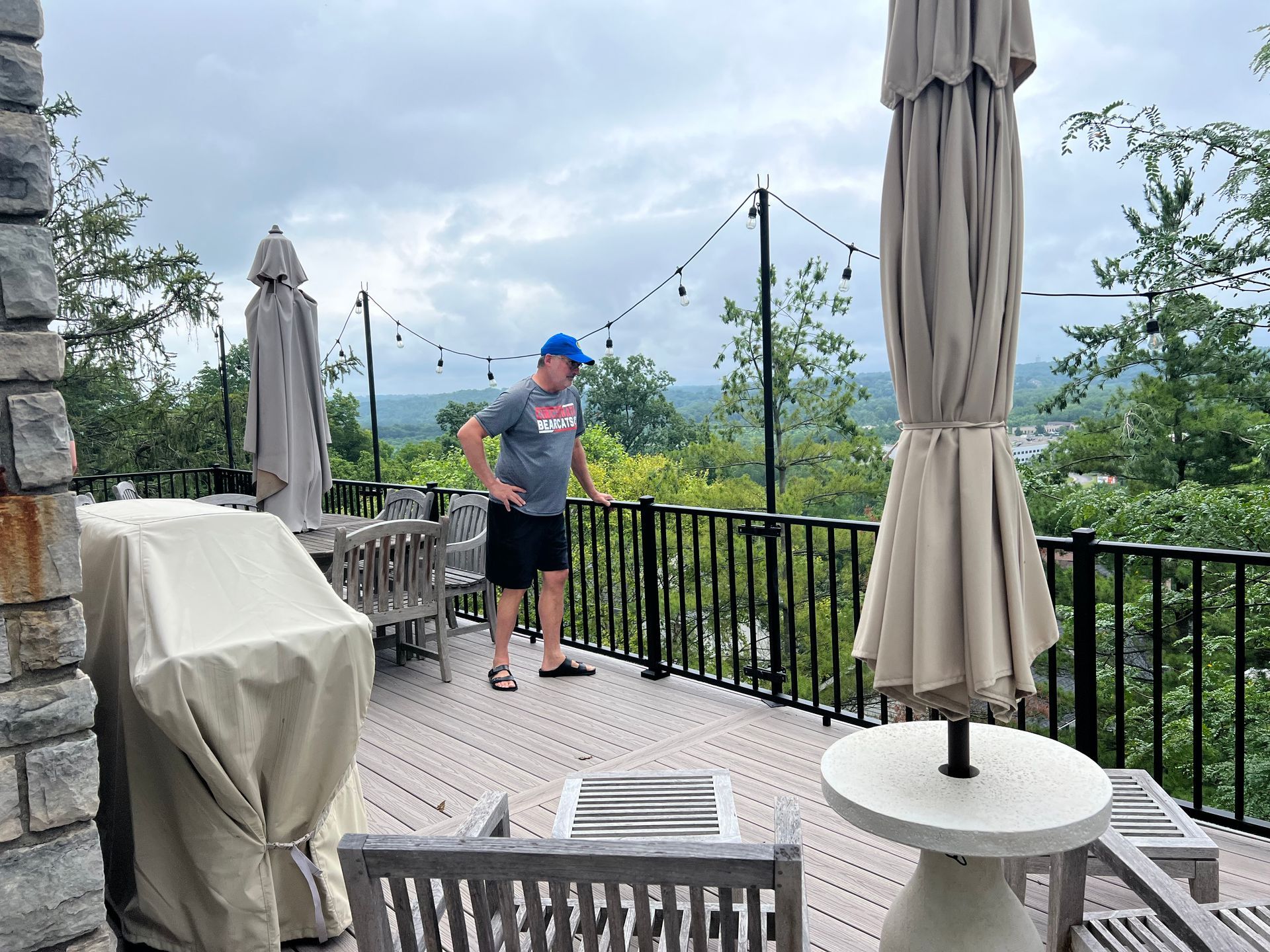 Person standing on a patio by a black railing, overlooking a cloudy mountain view with covered tables and umbrellas.