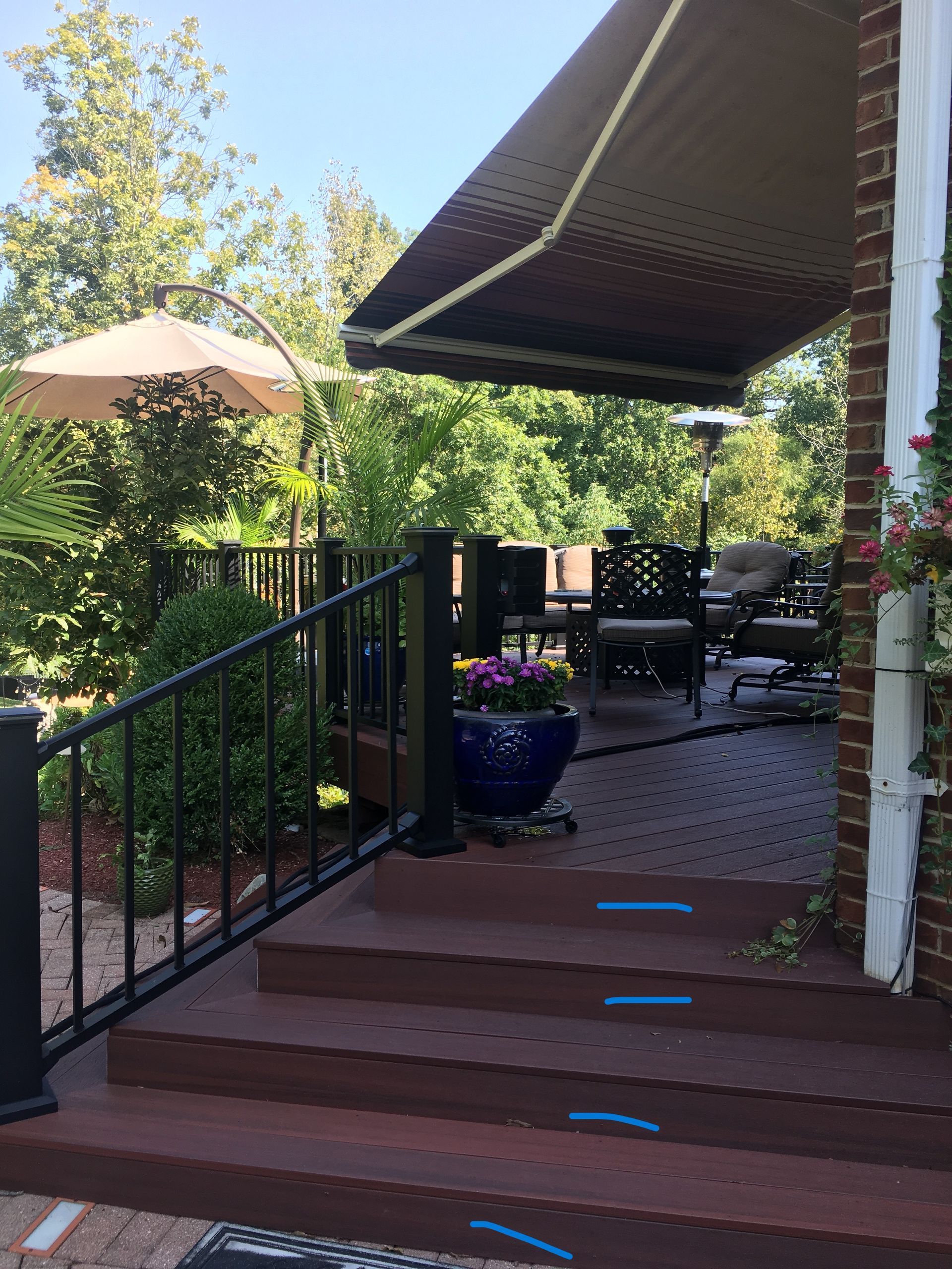 Backyard deck with steps, black railing, potted flowers, patio furniture, and an awning in sunlight