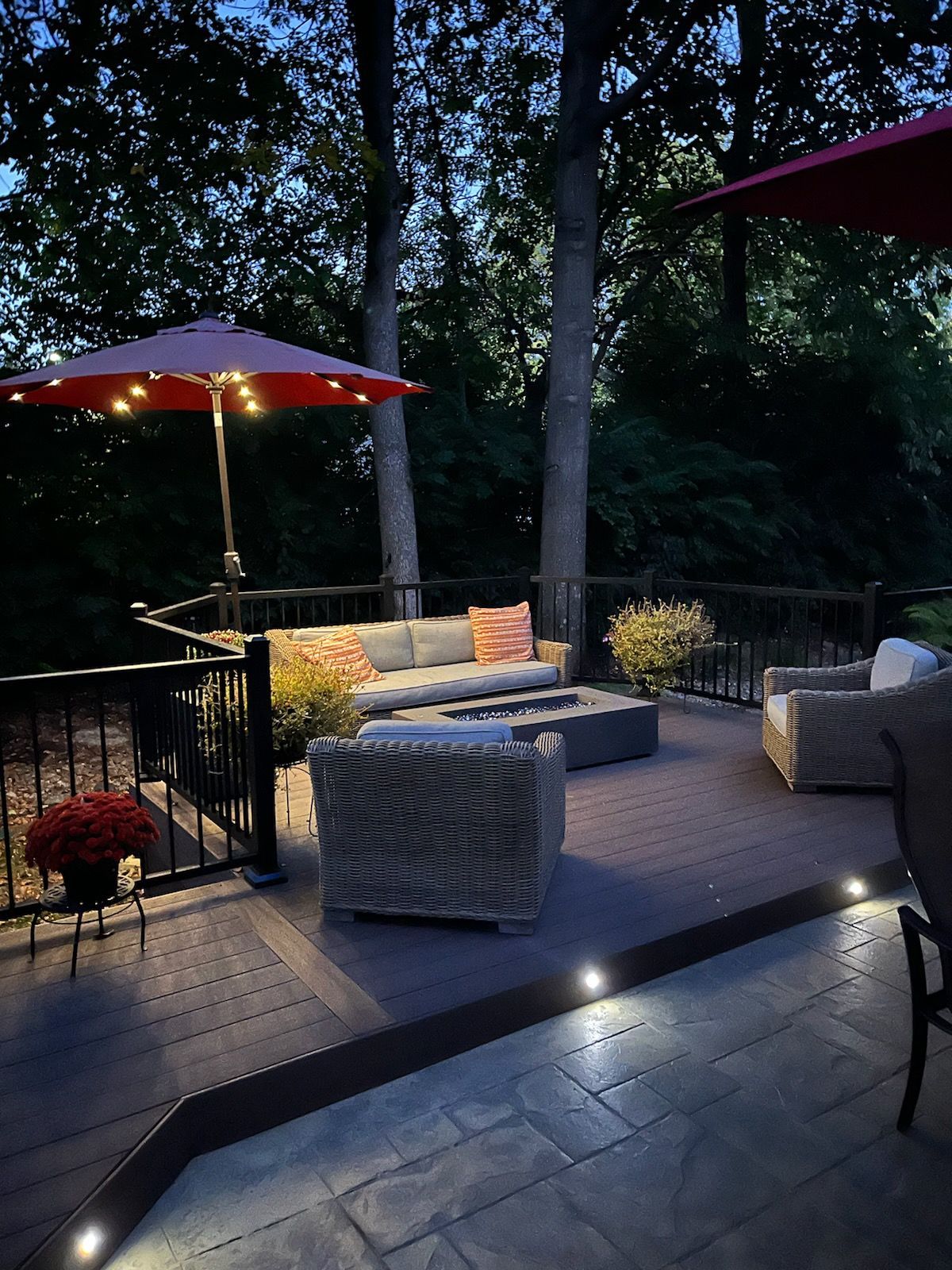 Outdoor patio seating area at dusk with cushioned chairs, red umbrellas, and soft pathway lights.