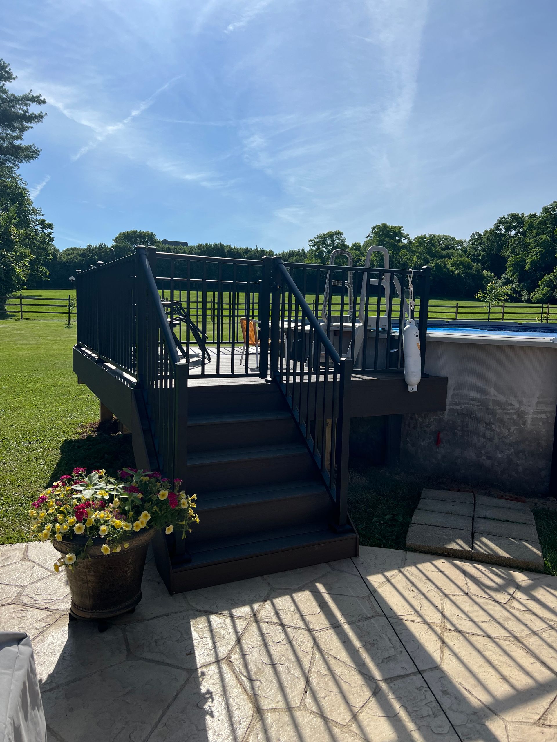 Black-railed deck steps beside a stone wall, with flower pots and a bright grassy field beyond.