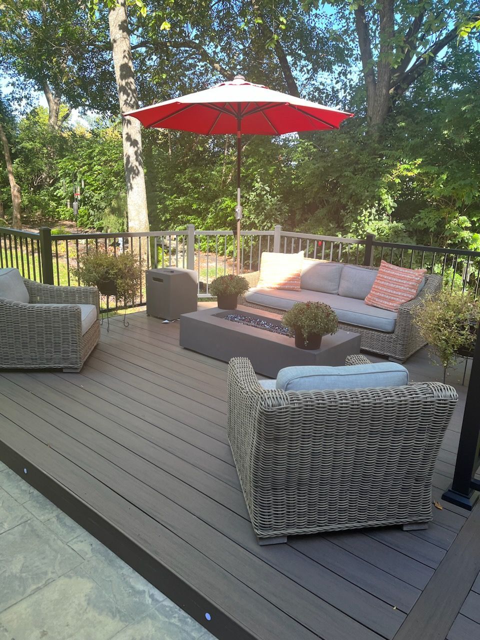 Outdoor deck seating area with wicker chairs, gray sofa, and red umbrella amid trees