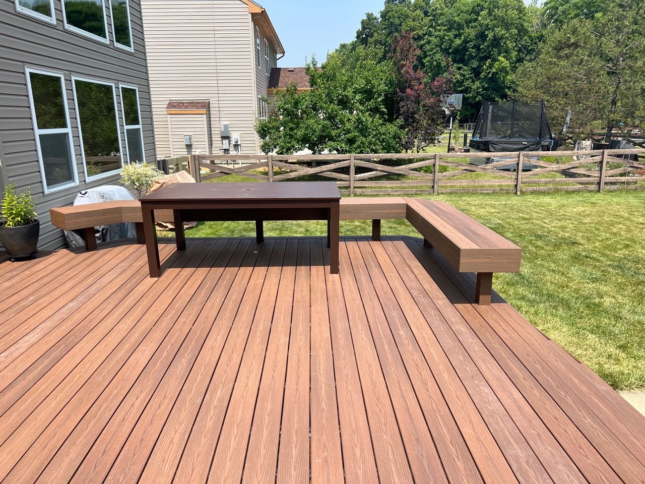 Backyard deck with wooden picnic table and bench beside a house, overlooking a lawn and trees