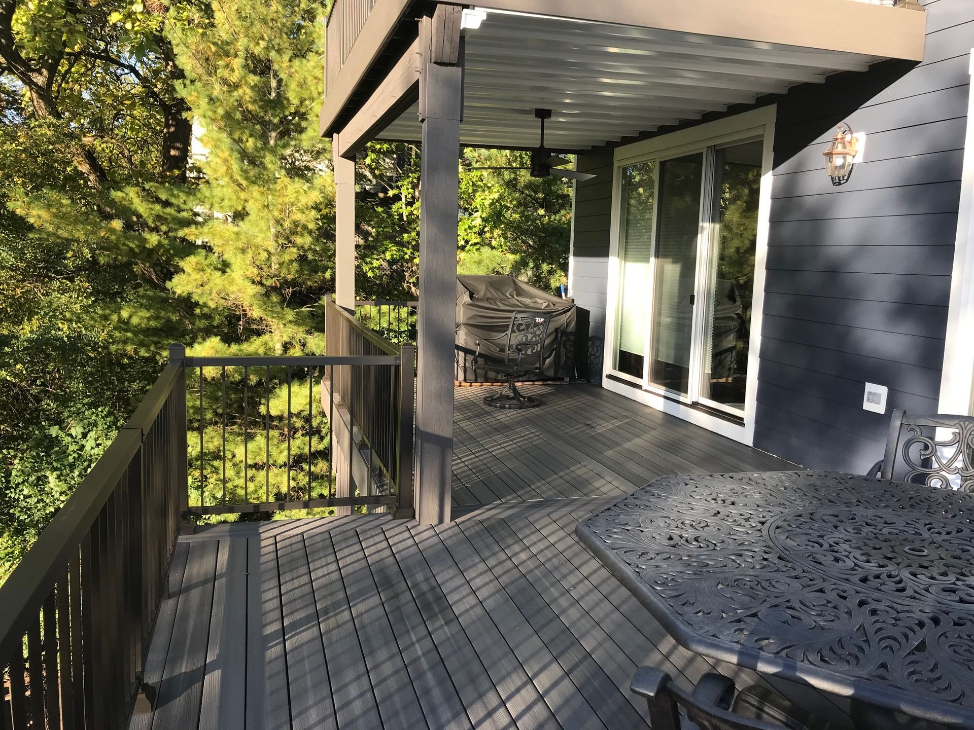 Wooden deck with black railing, metal patio table, sliding glass doors, and trees beyond the railing.
