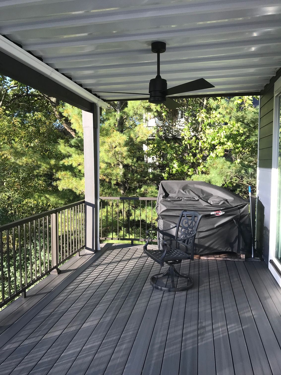 Covered porch with gray floorboards, black railing, fan, and a covered grill overlooking trees.