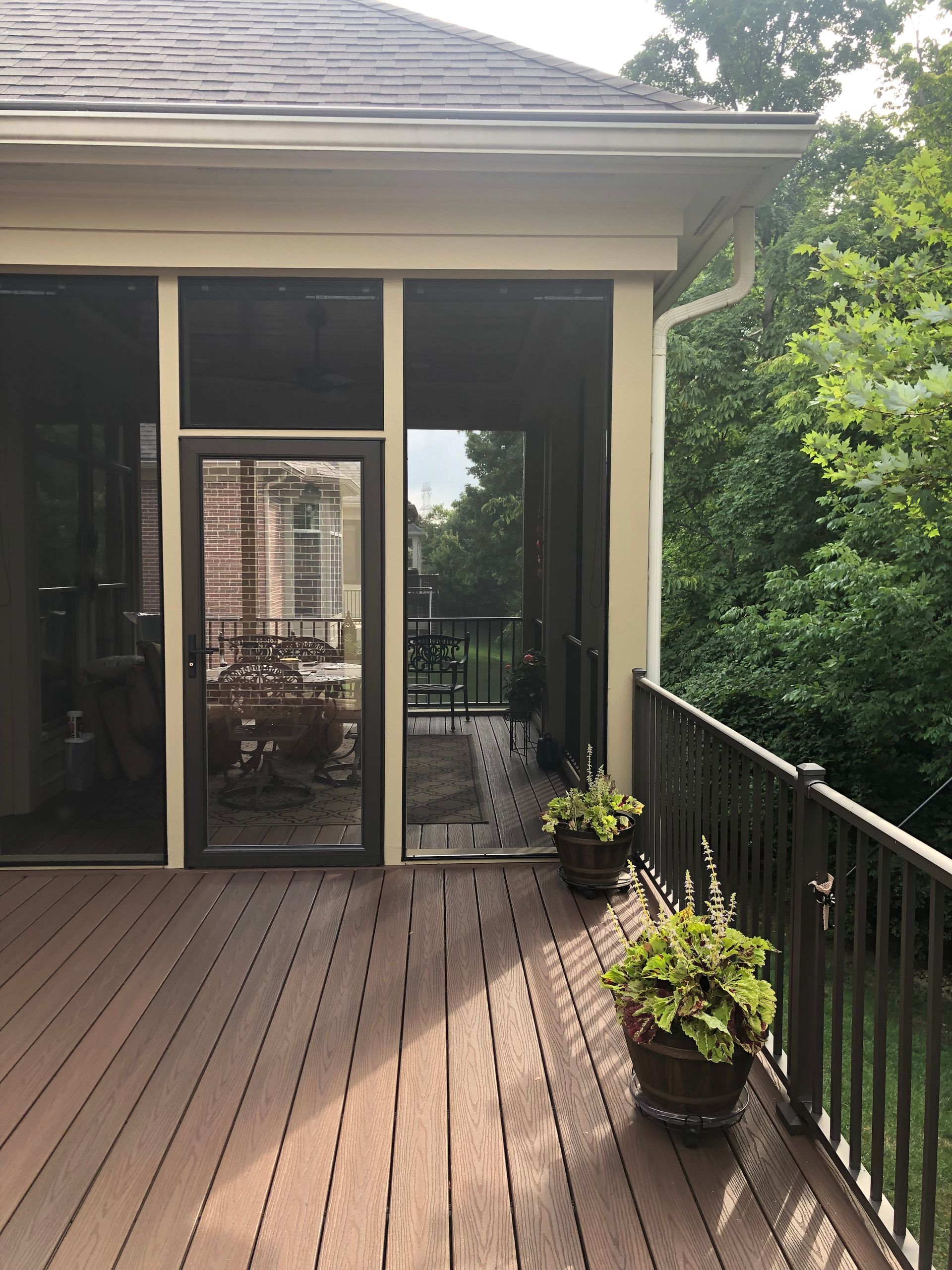 Wooden deck with potted plants beside a screened patio and black railing, with trees beyond.