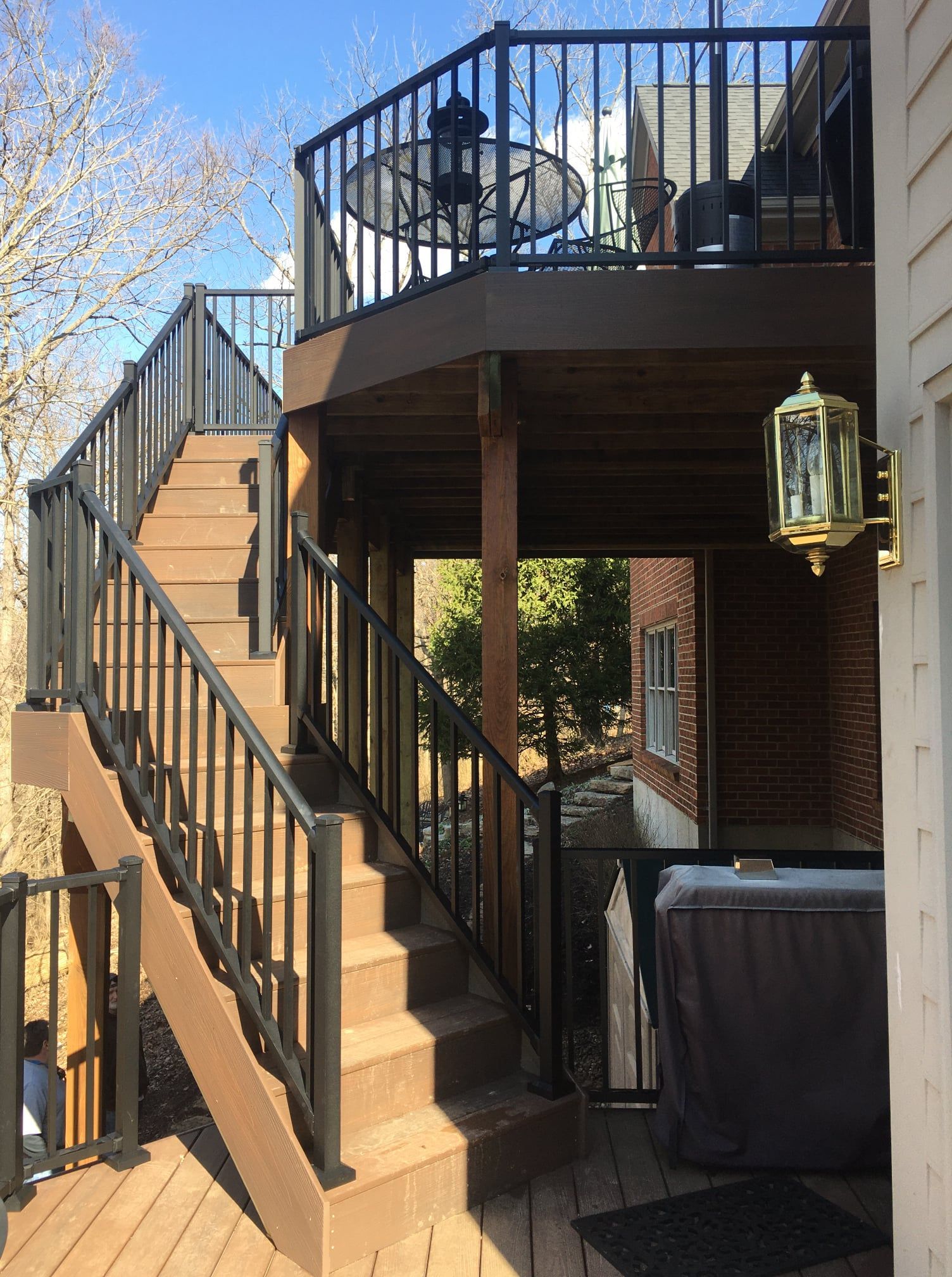 A wood deck with stairs and black railings leading up to an elevated balcony, viewed from the side of a house.
