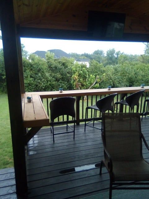 A covered deck featuring a wooden bar-style counter overlooking a grassy yard and dense green trees.