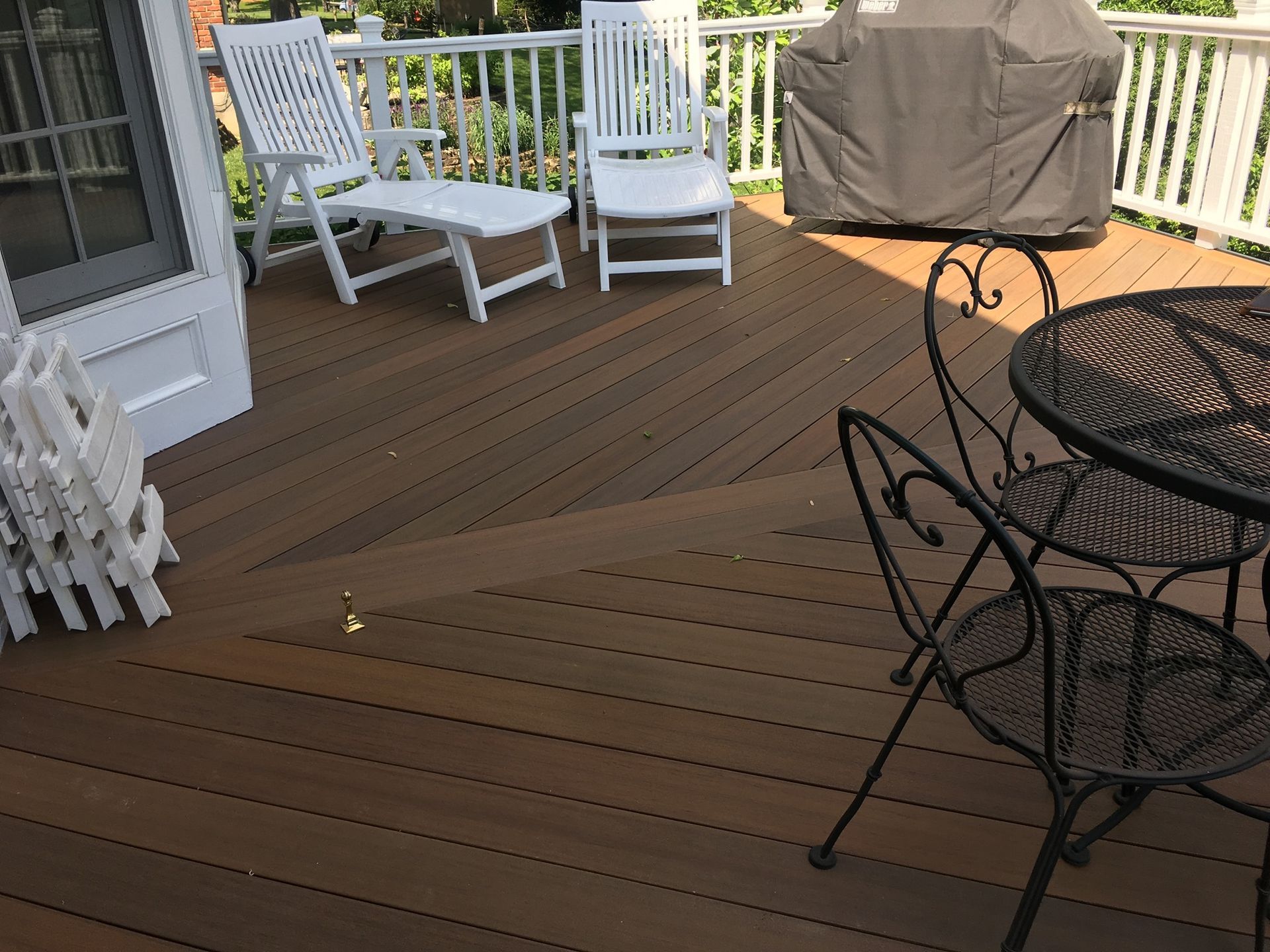 A wooden deck with white chairs, a small table set, and a covered grill under a railing.