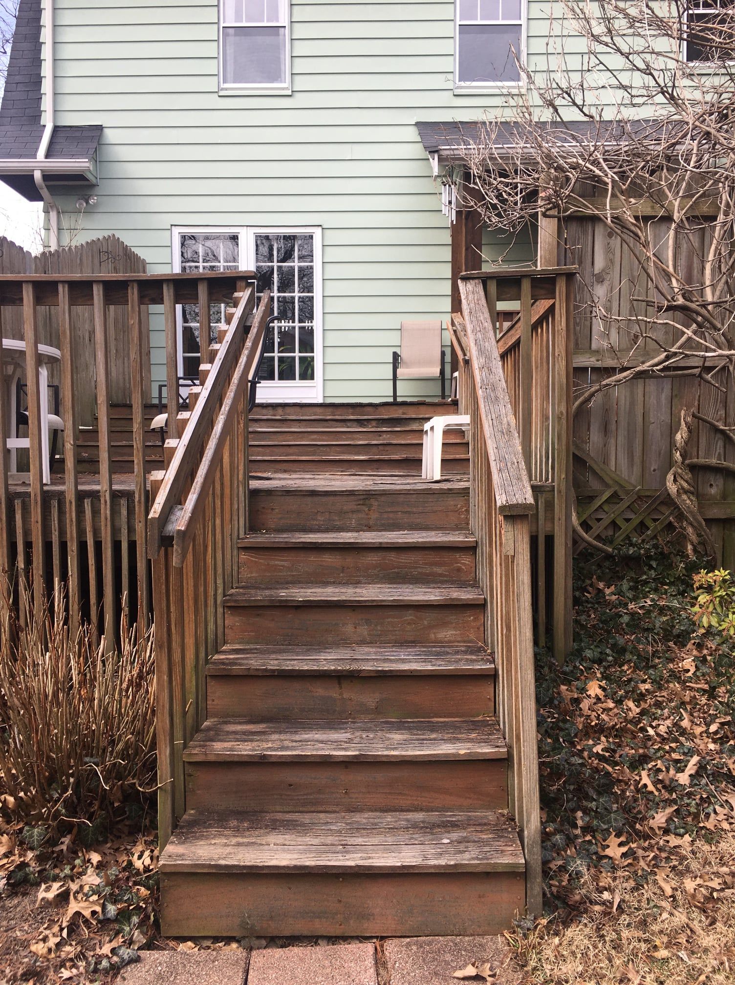 A set of weathered wooden stairs with handrails leading up to the back door of a light green house with a wooden deck.