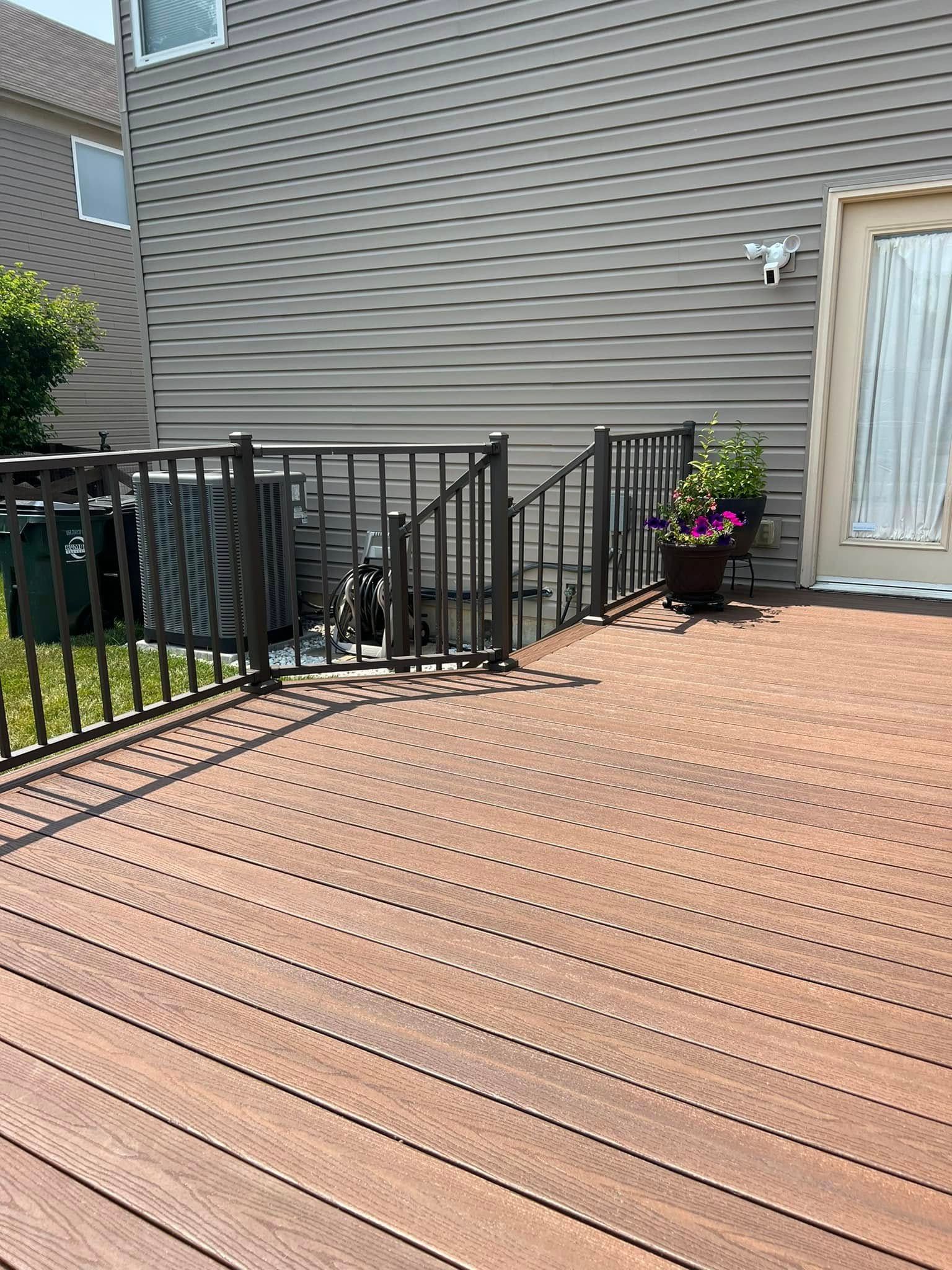 A tan-colored composite deck featuring a black metal railing, a side door, and a potted plant near the stairs.