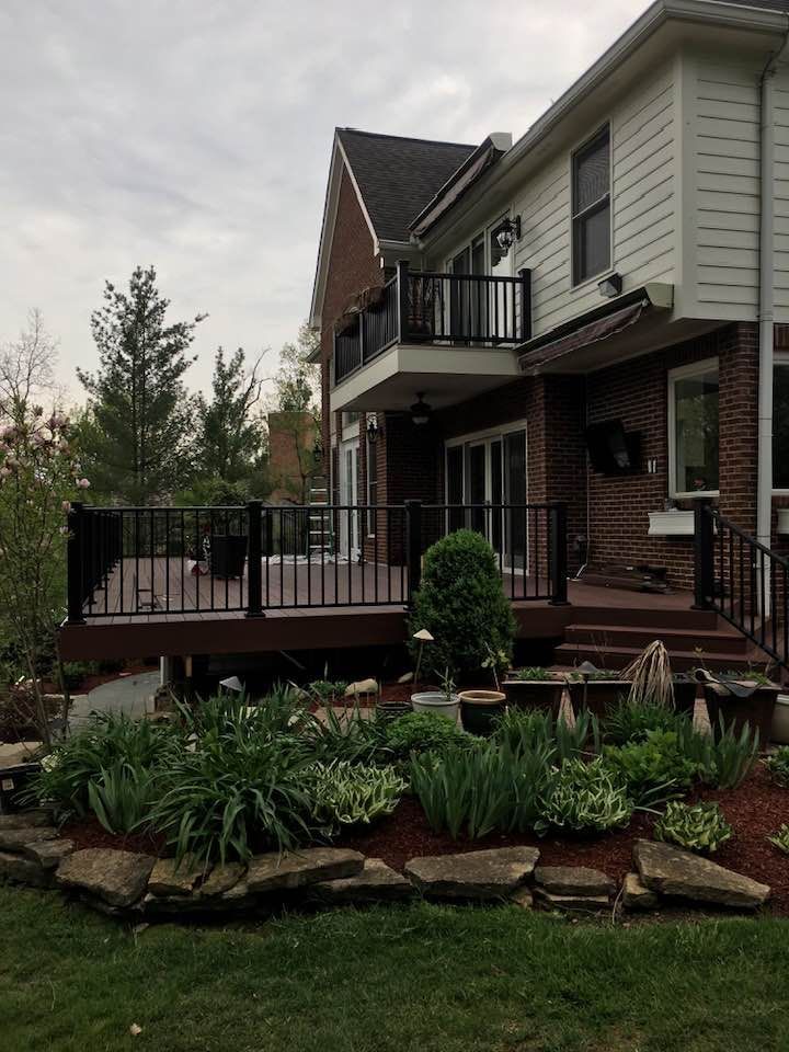 A brick and white-sided house with a large wooden deck, black railings, and a landscaped stone garden in the foreground.