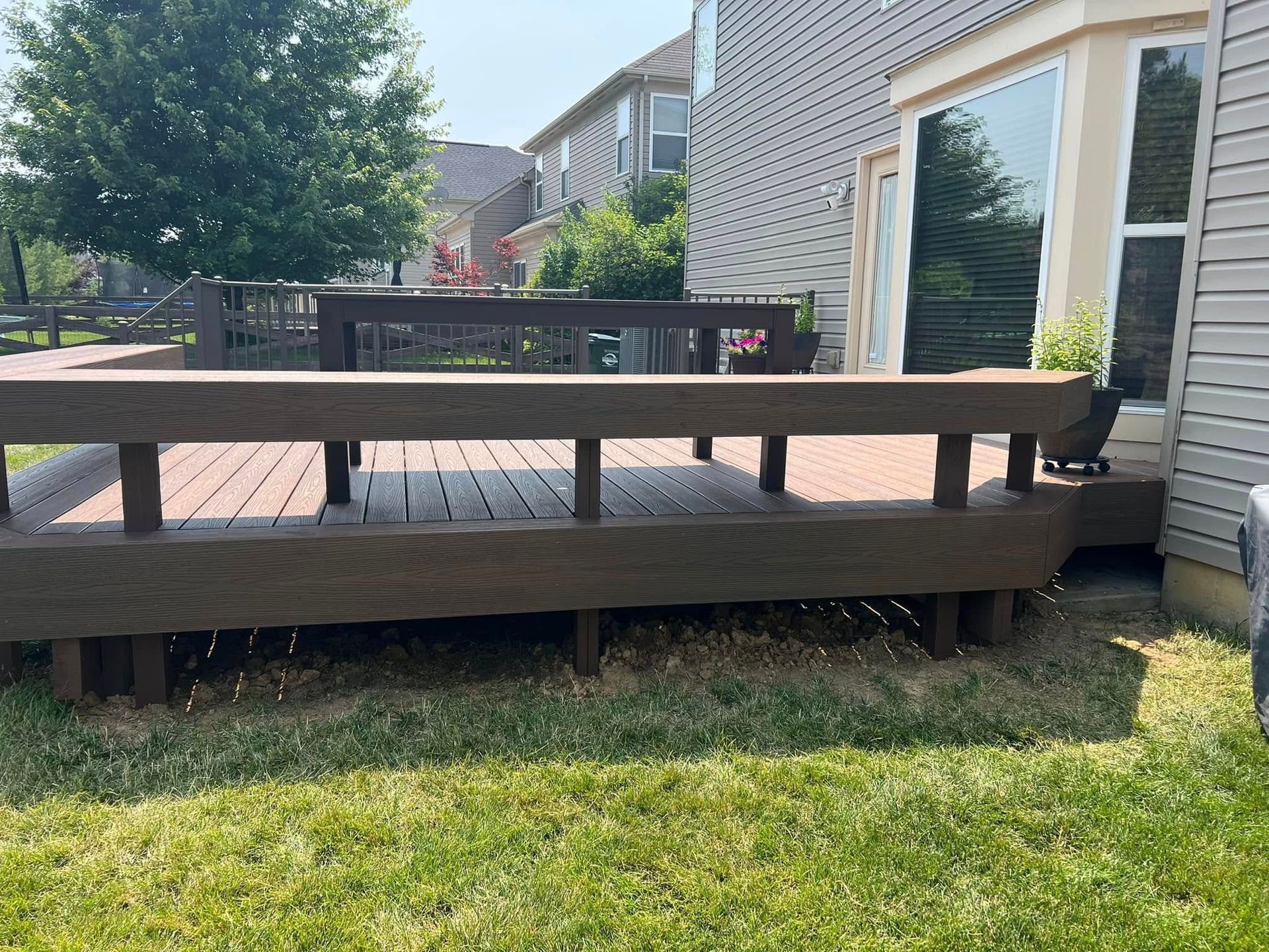 A dark wood deck with a wide railing extends from a residential house into a grassy backyard on a sunny day.
