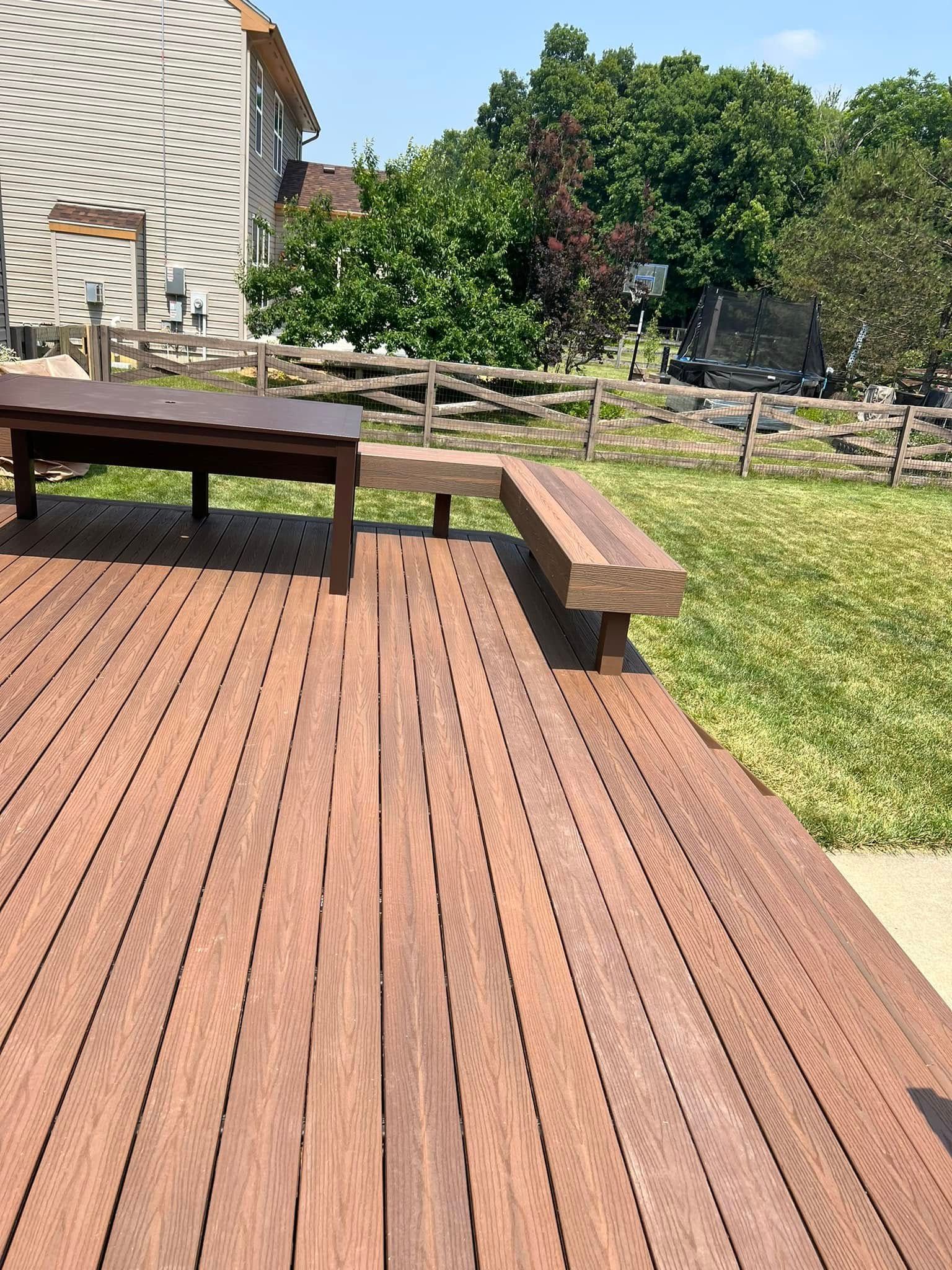 A wooden deck with a built-in corner bench and a small table, overlooking a grassy yard with a split-rail fence.