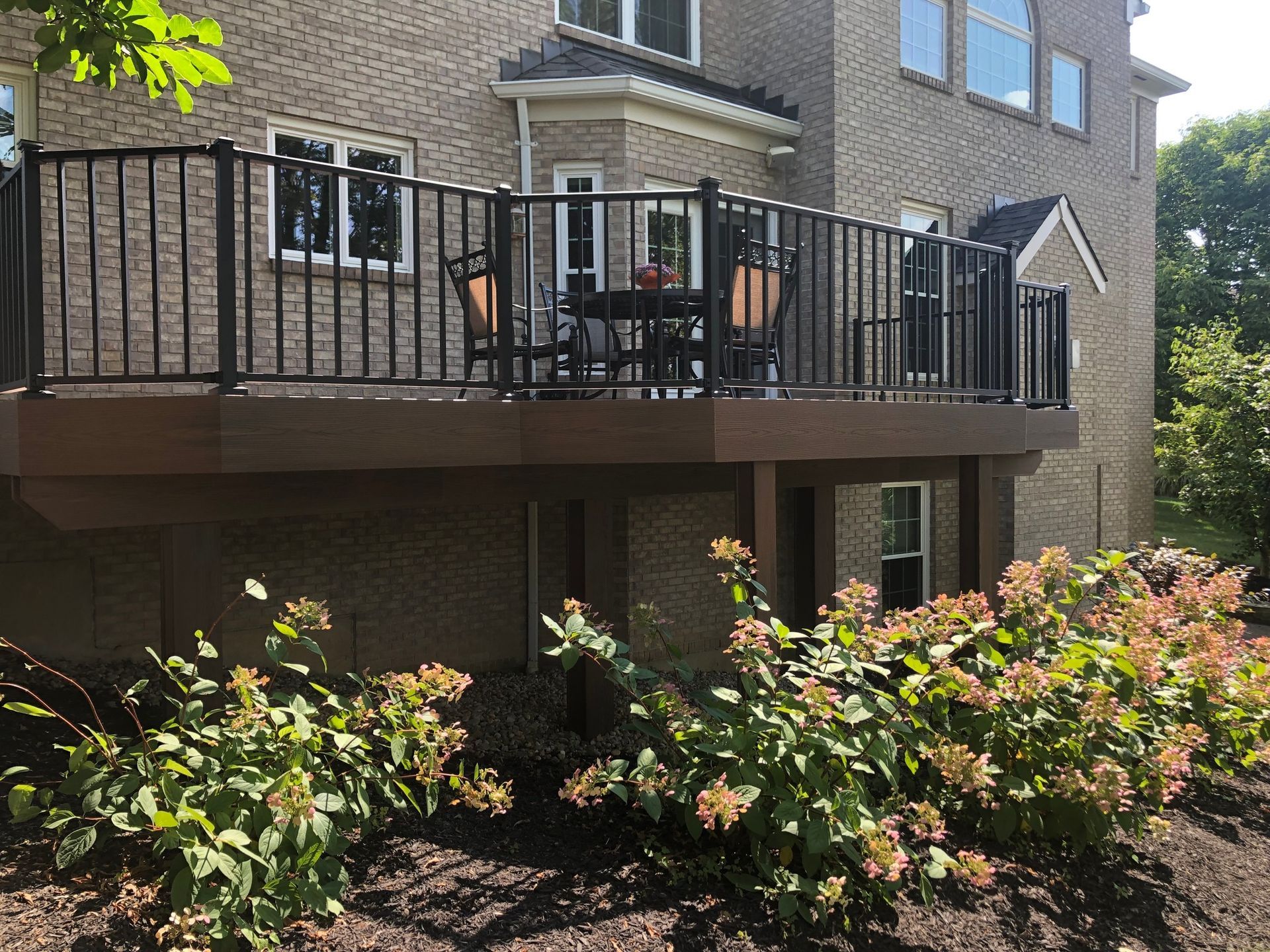 A raised composite deck with black railings and patio furniture attached to the rear of a multi-story brick house.