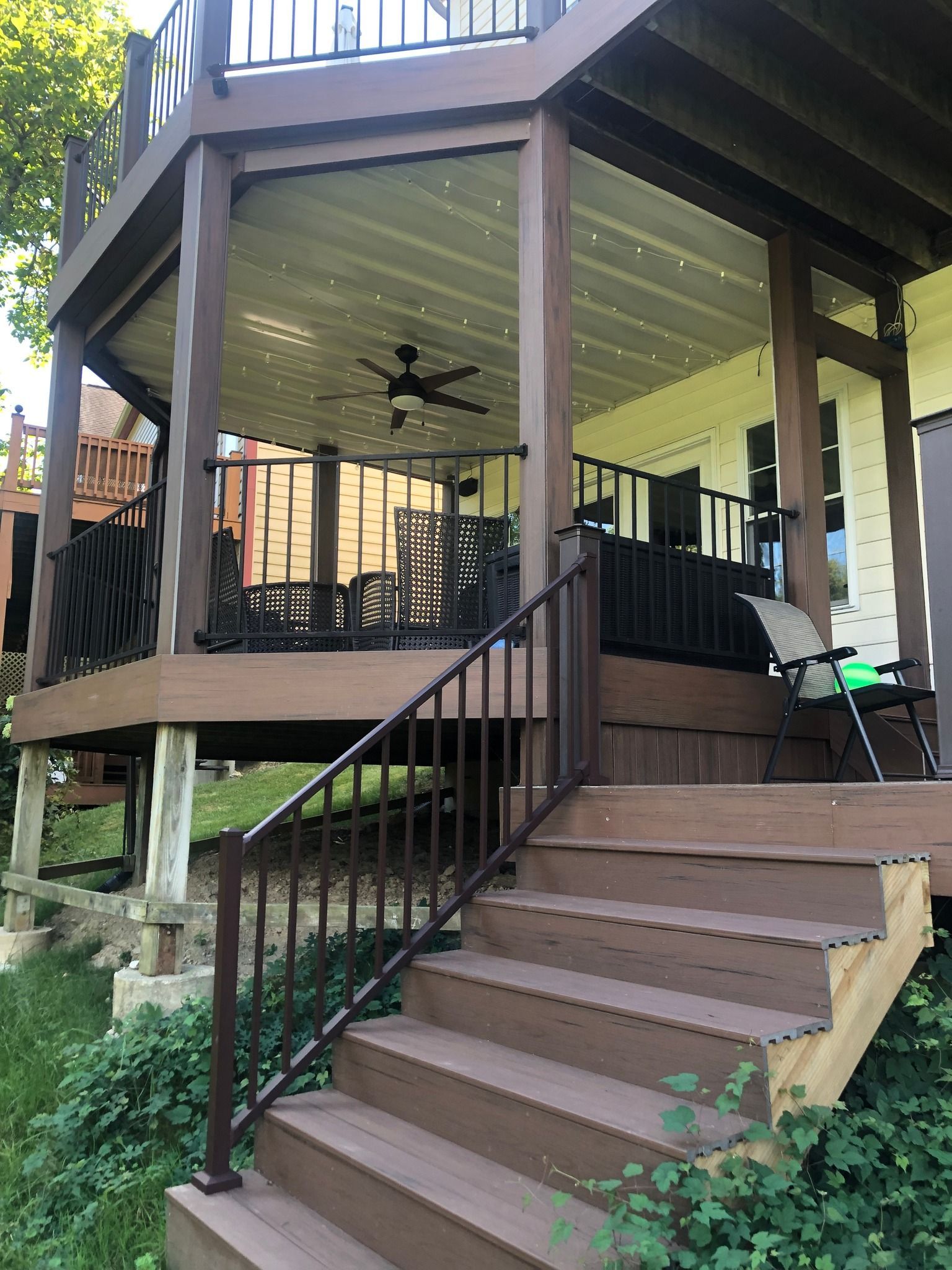 A brown wooden deck with stairs leading up, featuring a covered patio area with a ceiling fan and black railings.