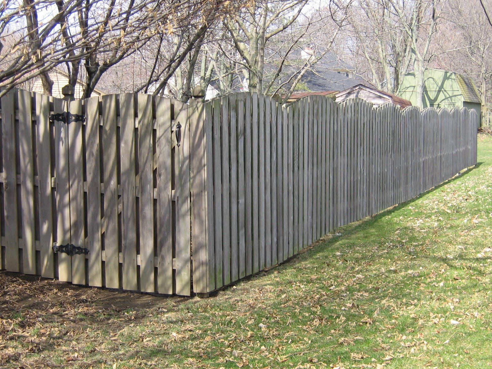 A weathered wooden picket fence with a scalloped top edge stands in a grassy yard on a sunny day.