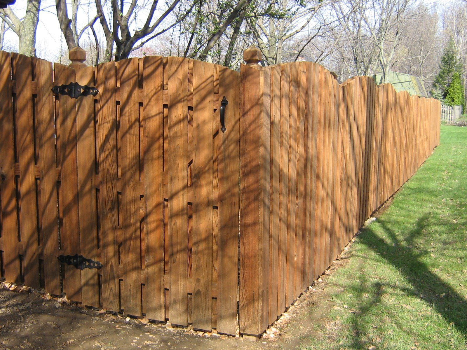 A brown, scalloped-top wooden fence with a gate, hinges, and a post cap, set against a grassy yard with tree shadows.