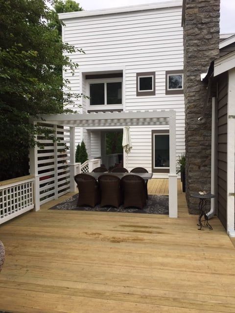 A wooden deck with a white pergola covering a dining set, adjacent to a white house and a stone chimney.