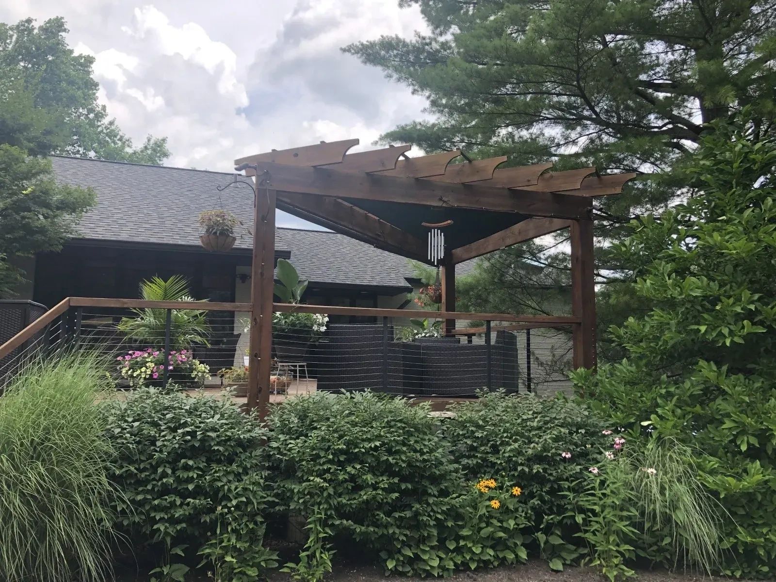 A wooden pergola on a backyard deck, surrounded by lush green shrubs and trees, with the house visible in the background.
