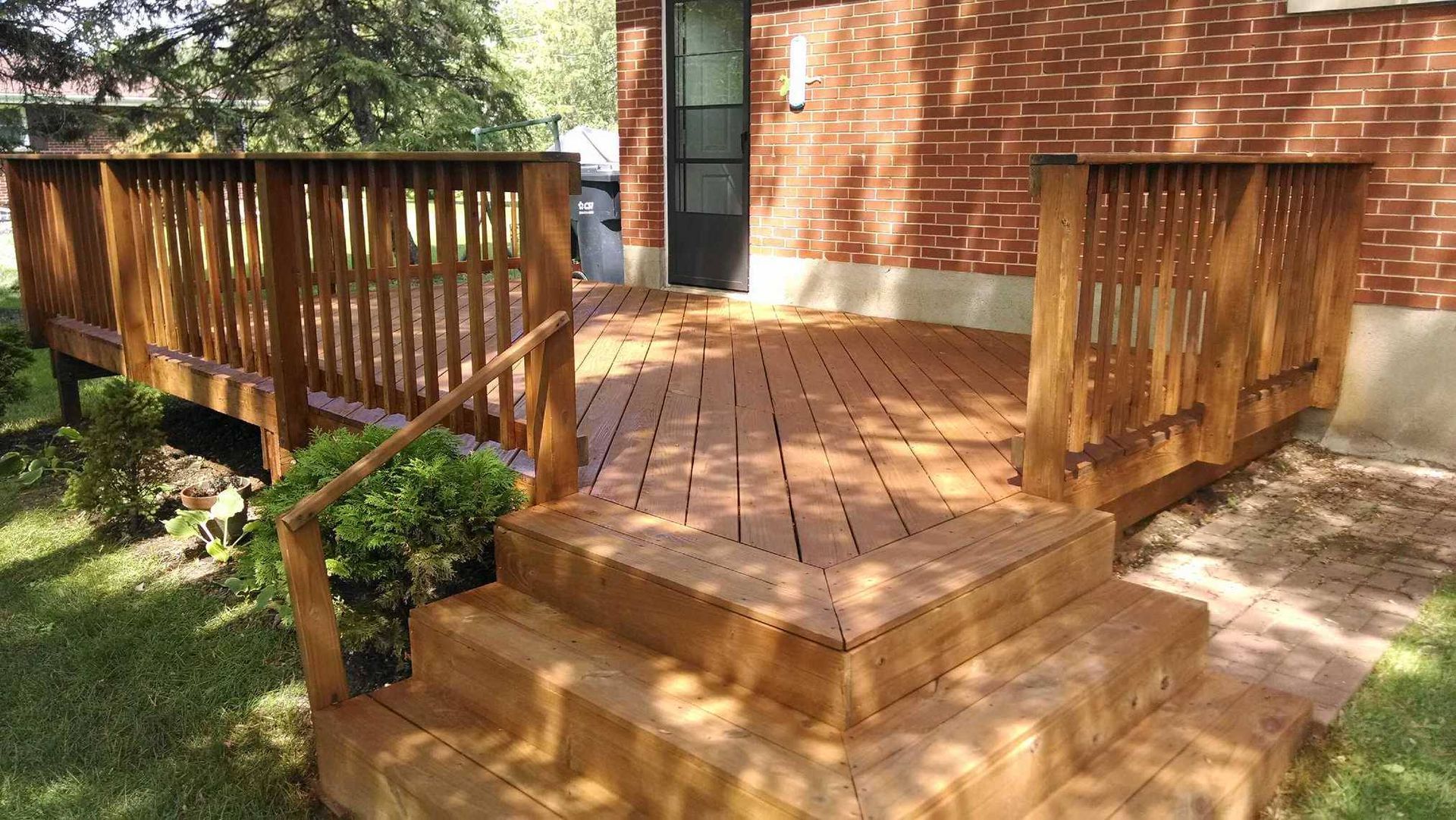 A wooden deck with a diagonal slat pattern, two steps leading down, and railings, attached to a brick house exterior.