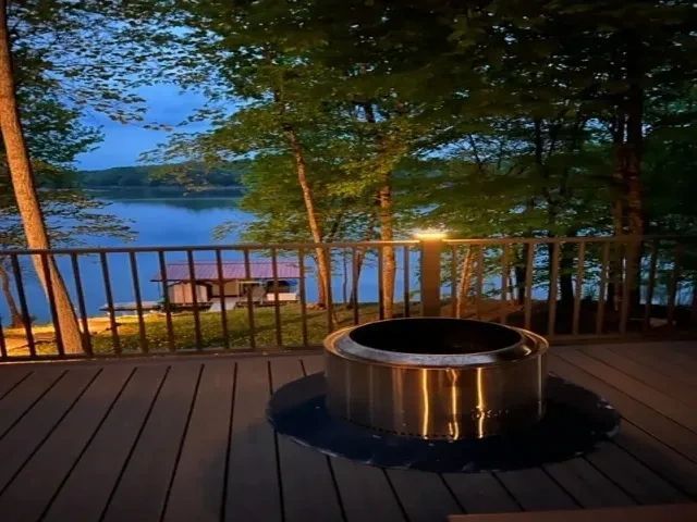 A stainless steel fire pit sits on a wooden deck overlooking a lake at dusk, framed by trees and a boat dock.
