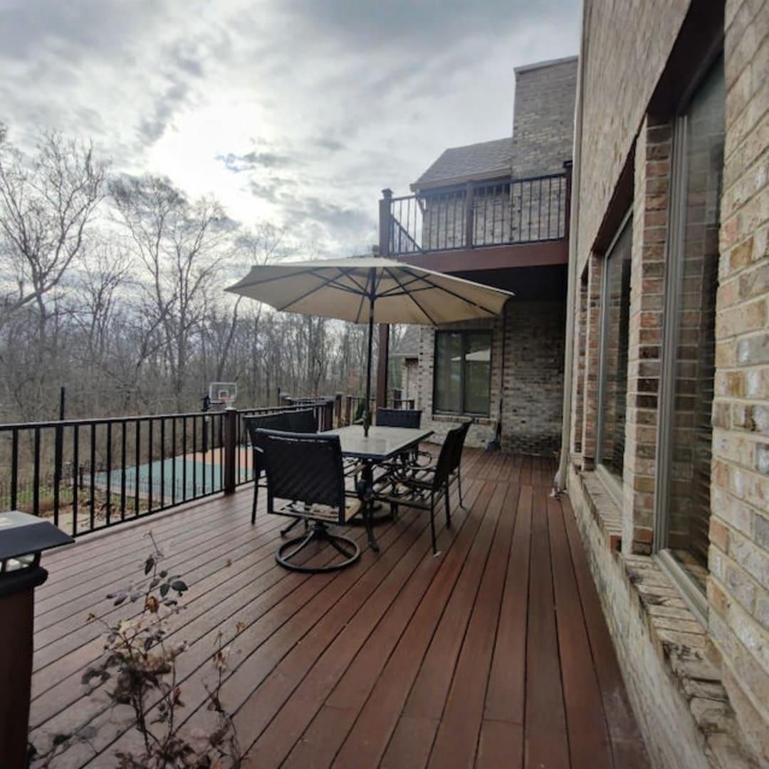 Stone house deck with patio table, umbrella, chairs, and wooded view on a cloudy day