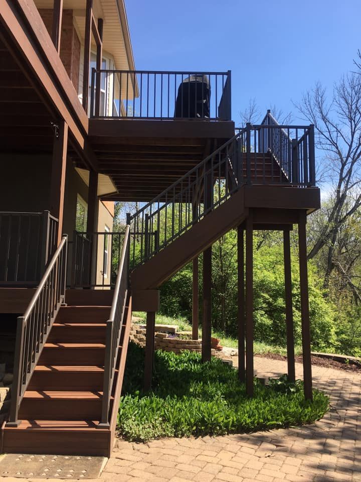 Multi-level wooden deck with dark brown railings and stairs leading from a paved patio up to the house.