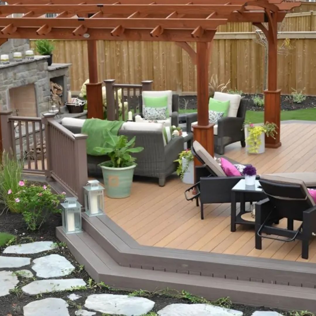 A tiered wooden deck with a pergola, stone fireplace, outdoor seating, and greenery, set in a backyard.