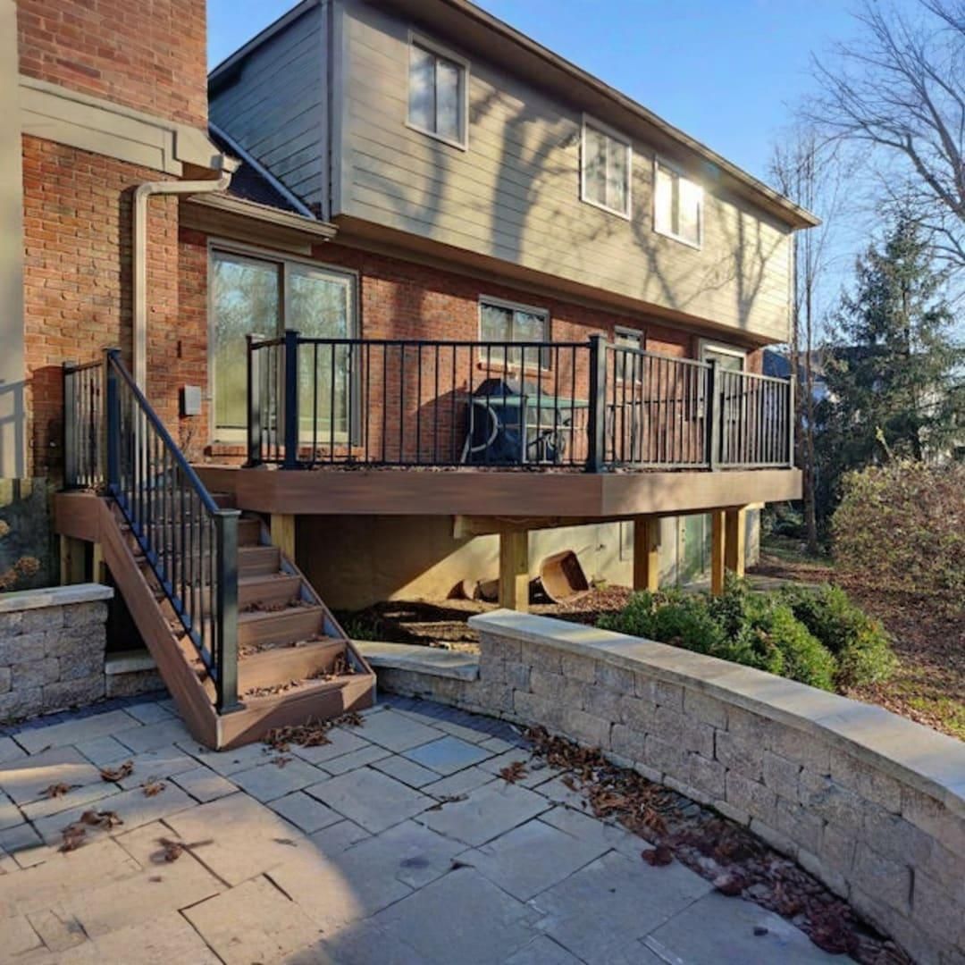 A wooden deck with black railings and stairs overlooks a paved stone patio and retaining wall by a brick and siding house.