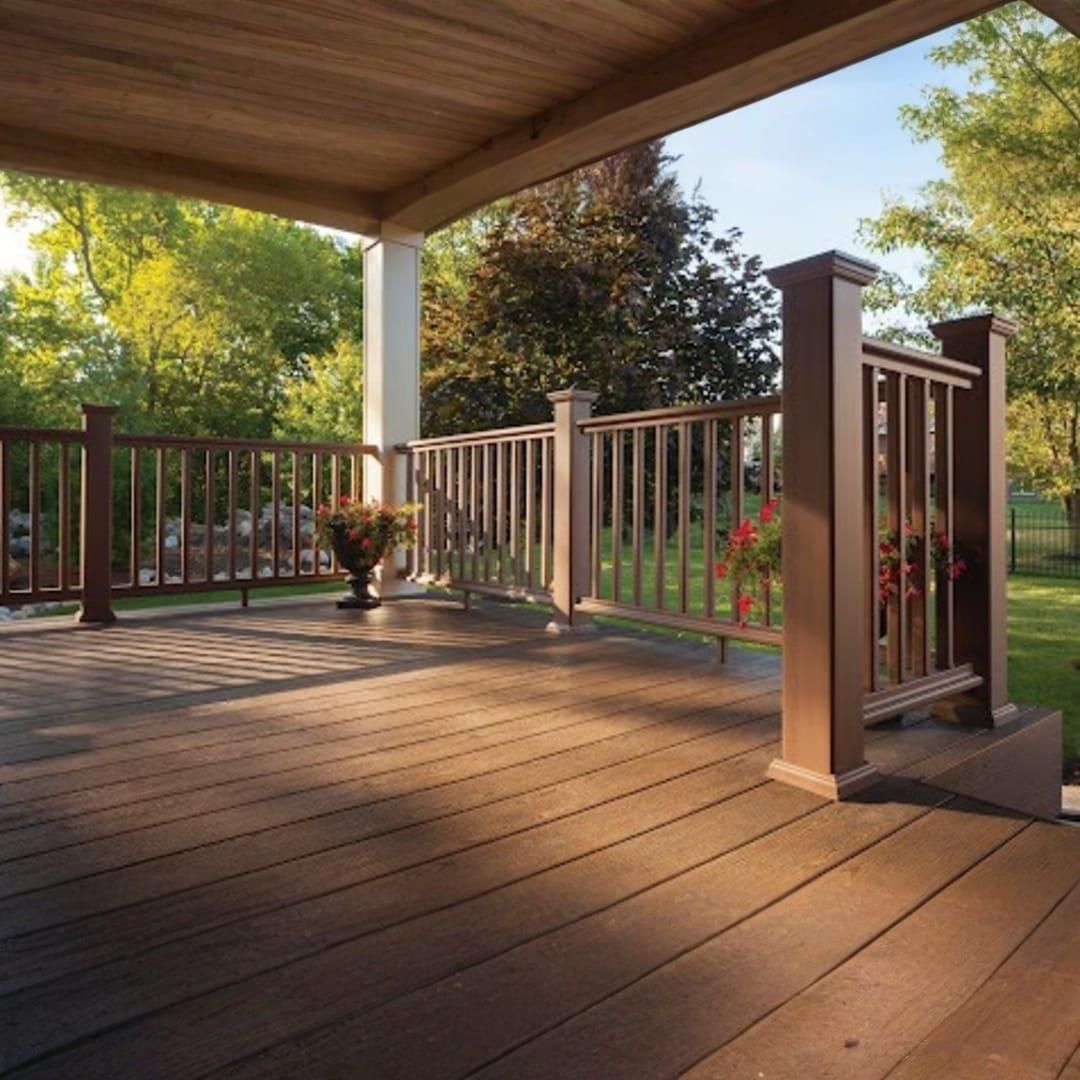 A covered wooden deck with a brown railing, overlooking a green yard with trees.