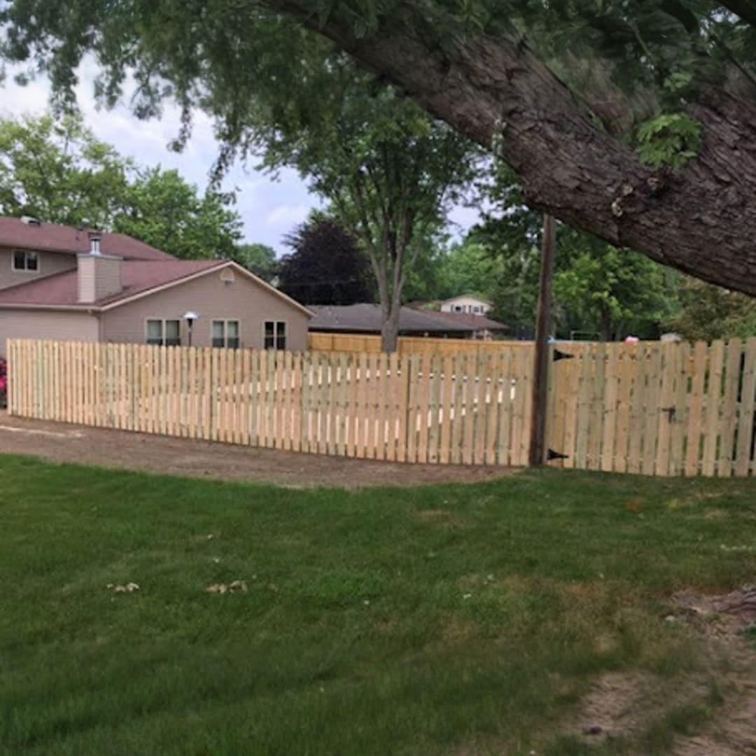 A new wooden picket fence encloses a backyard pool area on a grassy lawn near a residential house and a large tree.