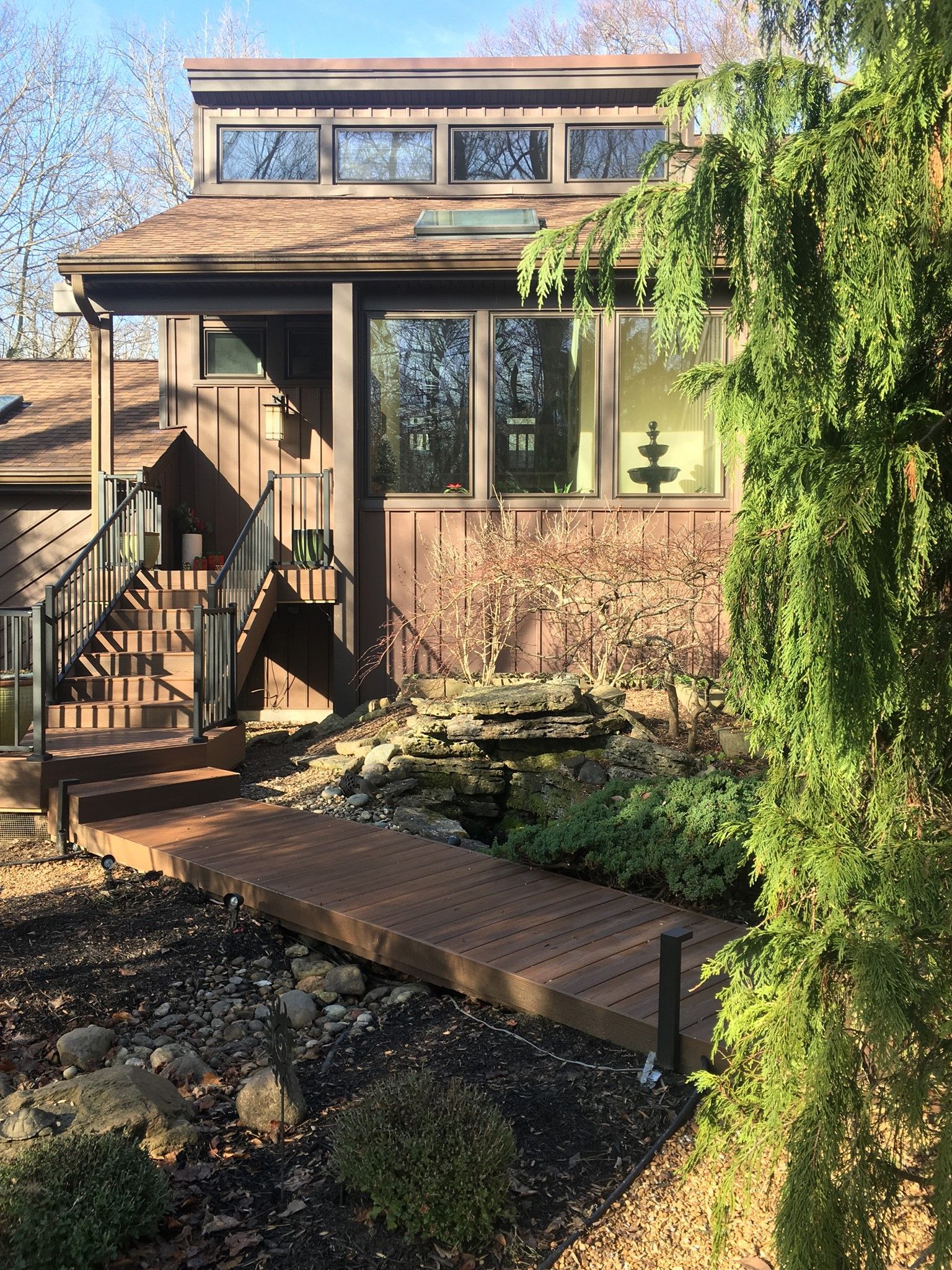A brown house with a prominent clerestory window, front stairs, and a wooden walkway leading to a stone garden feature.