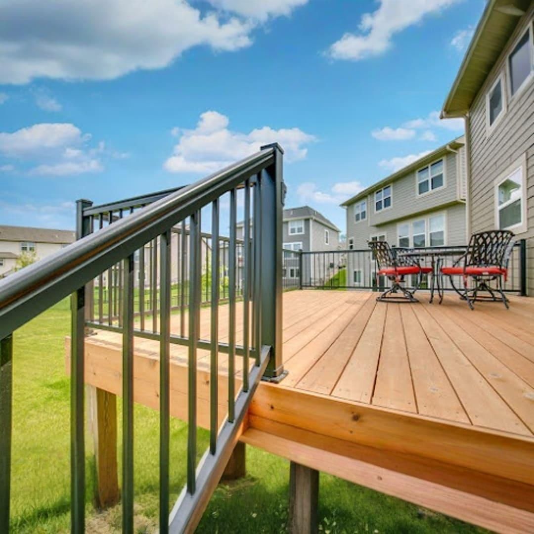 A wooden deck with black metal railings and patio furniture, overlooking a grassy backyard under a blue sky.